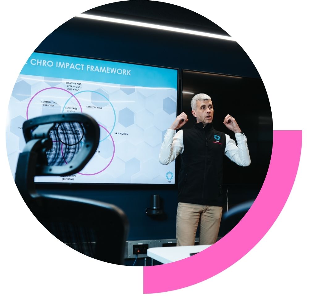 Tom Emery presenting at the front of a meeting room as part of his coaching programme and one day experience. With a big screen and the CHRO framework on the screen